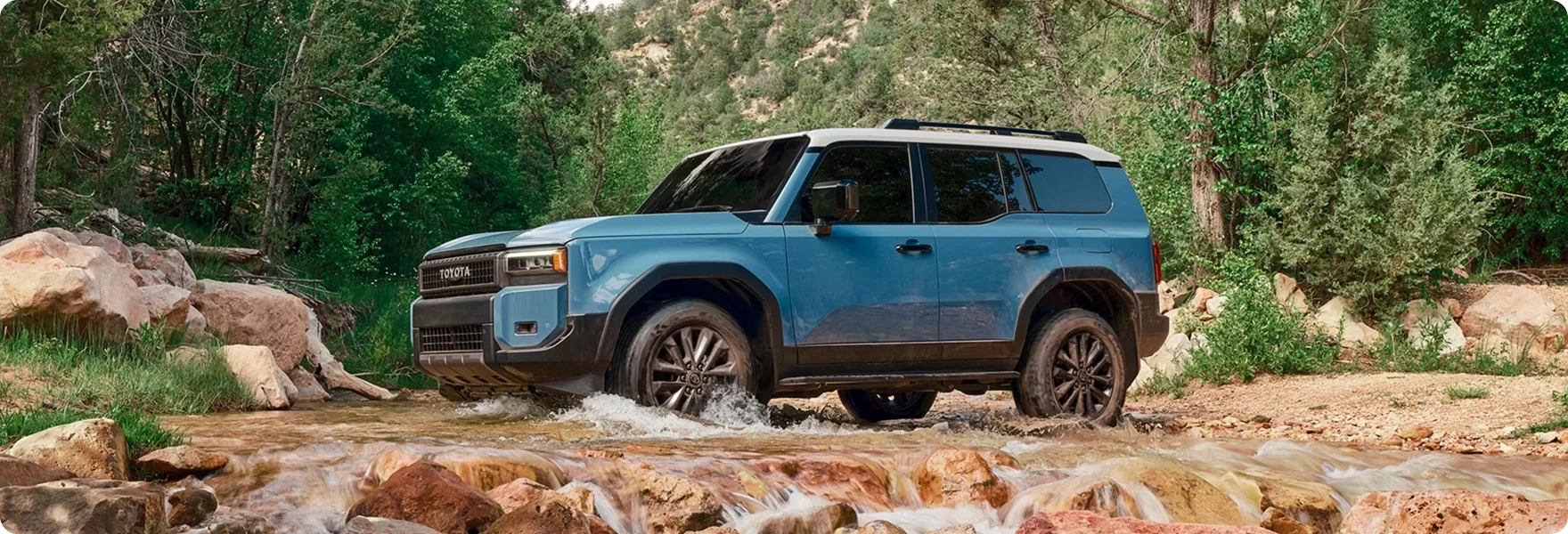 Blue 2027 Toyota Land Cruiser fording a shallow stream on a forest trail
