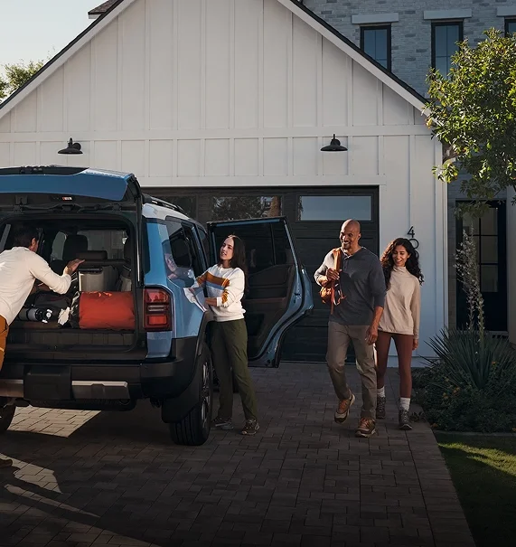 People loading gear into the open rear cargo area of a blue Land Cruiser outside a farmhouse