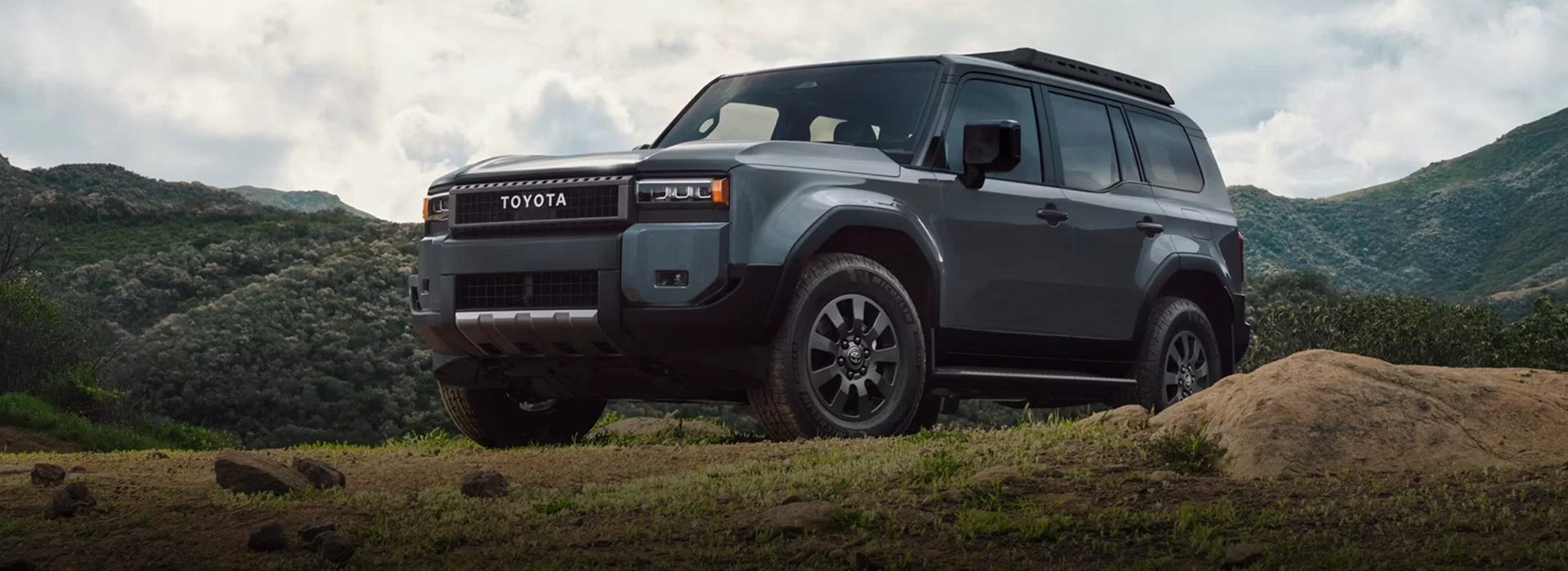 Slate-gray 2027 Toyota Land Cruiser parked on a rocky overlook with green mountains behind it