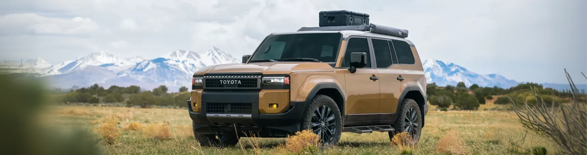 Bronze 2027 Toyota Land Cruiser with roof-rack gear in a meadow below snow-capped mountains