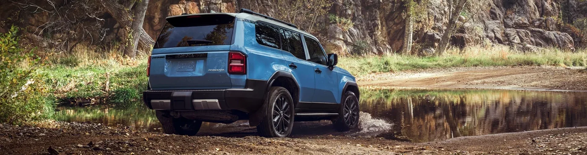 Blue 2027 Toyota Land Cruiser driving through a forest water crossing, rear view