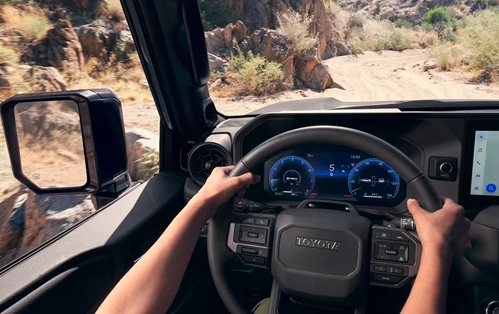 Driver&rsquo;s view of the Land Cruiser steering wheel and digital gauge cluster on a desert trail