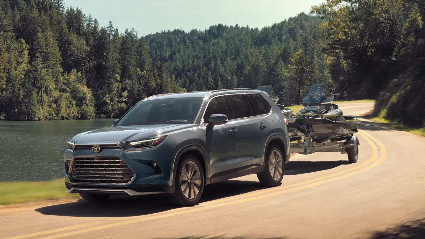Blue 2026 Toyota Grand Highlander towing jet-skis on a lakeside road — Dalton Toyota National City
