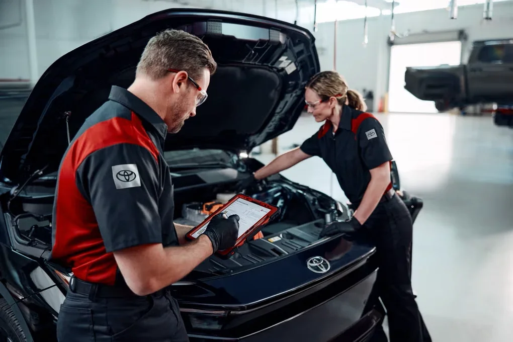 Two Toyota technicians at an open hood, one reading the inspection on a tablet