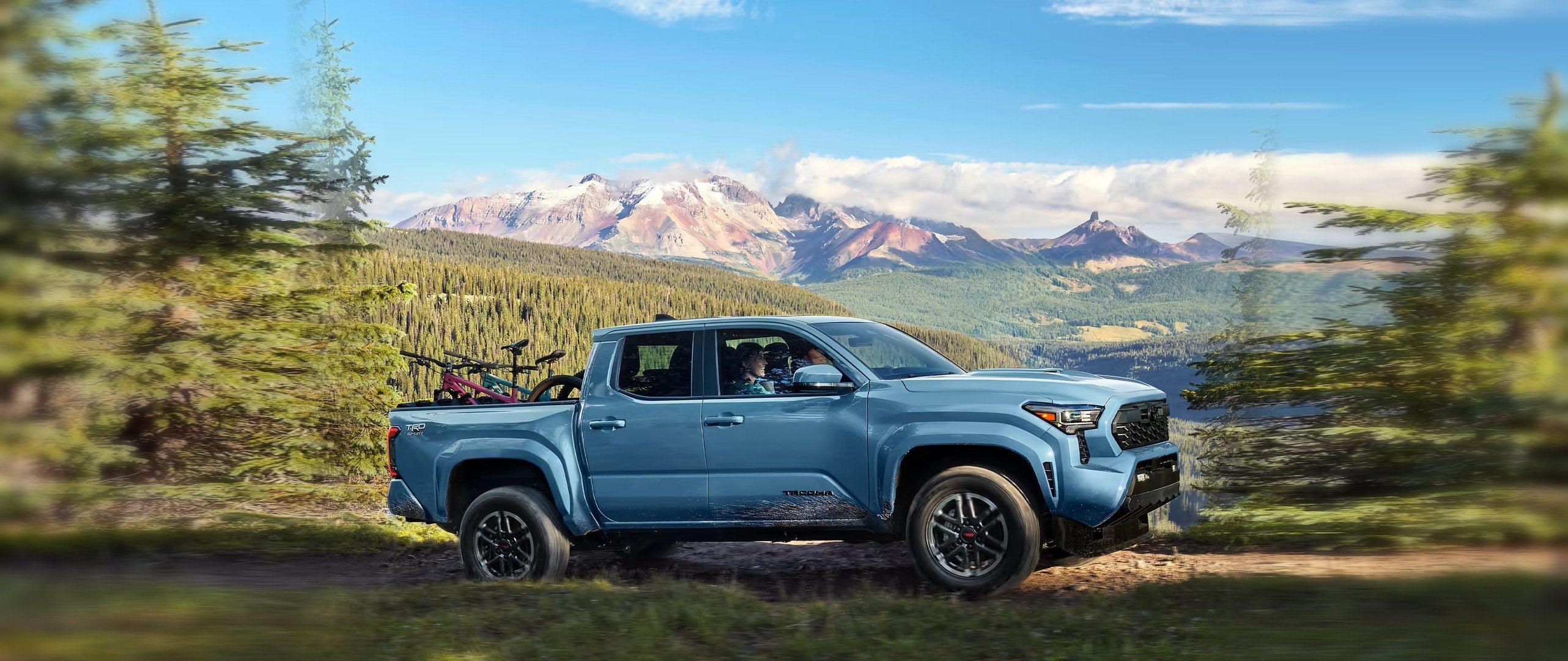 A blue Toyota Tacoma TRD Sport double cab with mountain bikes in the bed, parked on a forest trail with snow-capped mountains behind